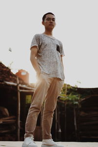 Low angle portrait of young man with hands in pockets standing on road against clear sky during sunset