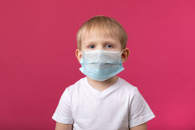 Child preschooler boy closeup shot in medical mask looking at the camera on a pink background. 