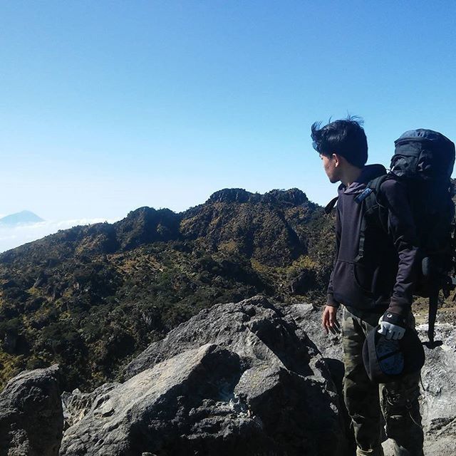 lifestyles, leisure activity, mountain, clear sky, men, casual clothing, hiking, standing, full length, rock - object, copy space, landscape, rear view, backpack, blue, tranquility, tranquil scene, nature