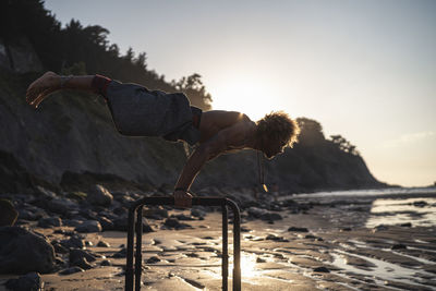 Shirtless young man exercising on parallel bars at beach against sky