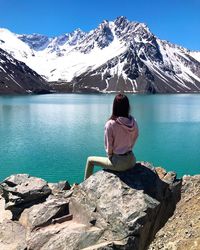 Rear view of man sitting on rock by lake against mountain