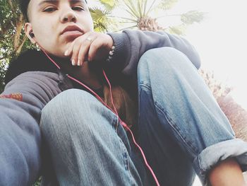 Low angle portrait of young man listening music while sitting by palm tree