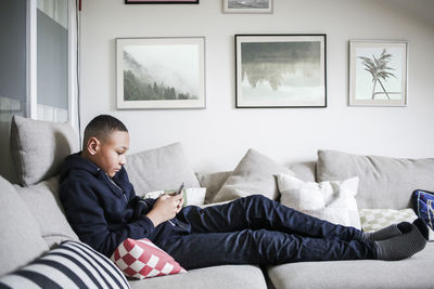 Full length side view of teenage boy using mobile phone on sofa at home