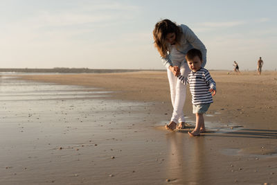 Full length mother assisting baby boy walking on shore at beach against sky