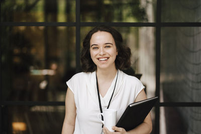 Portrait of cheerful young female entrepreneur standing with file and coffee cup in front of glass wall