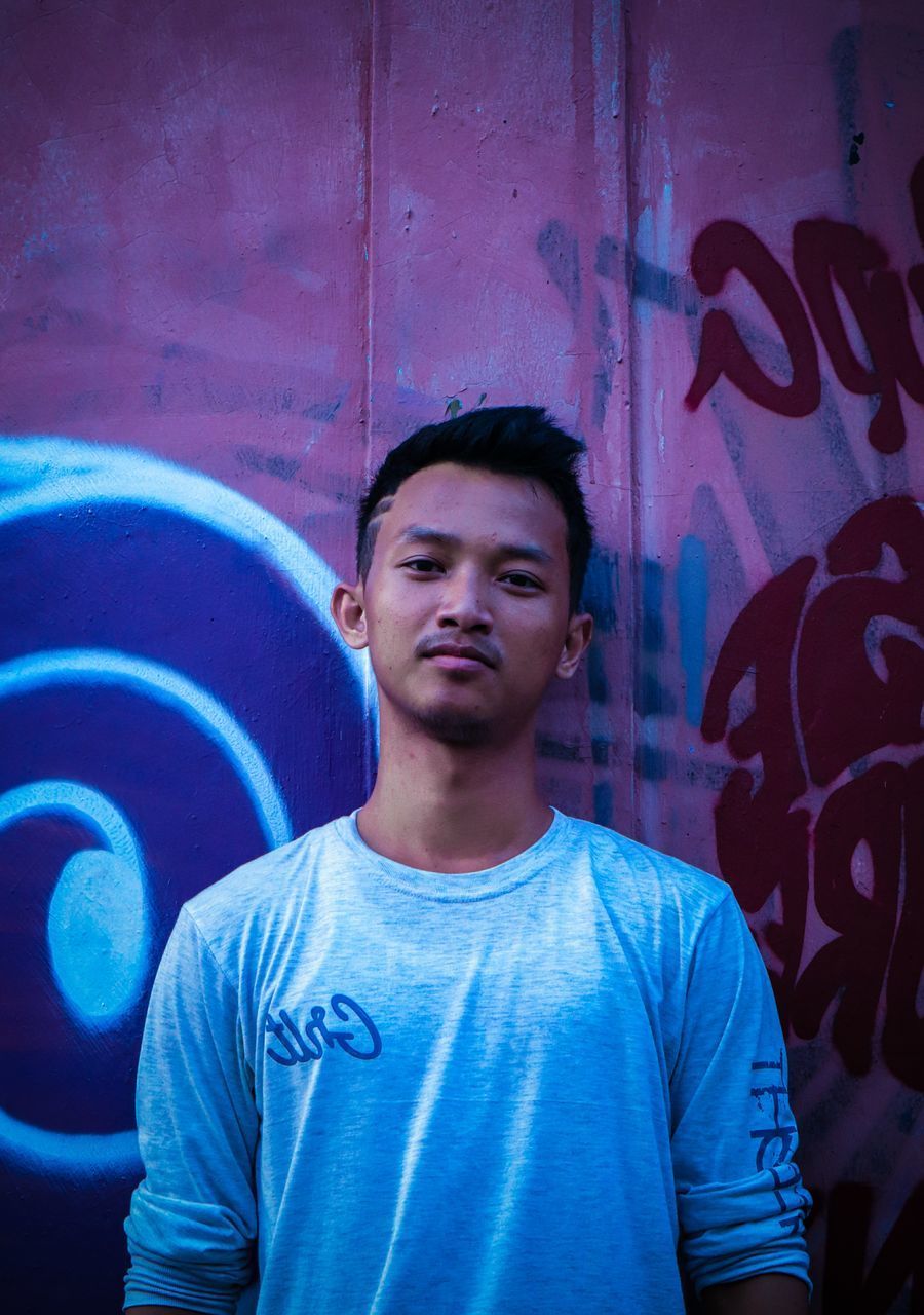 PORTRAIT OF HANDSOME YOUNG MAN STANDING AGAINST GRAFFITI WALL