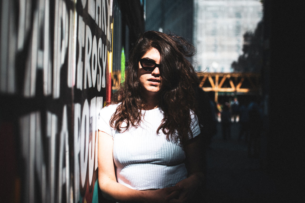PORTRAIT OF YOUNG WOMAN IN SUNGLASSES STANDING OUTDOORS