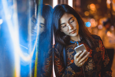 Smiling young woman using smart phone by window with reflection at night 