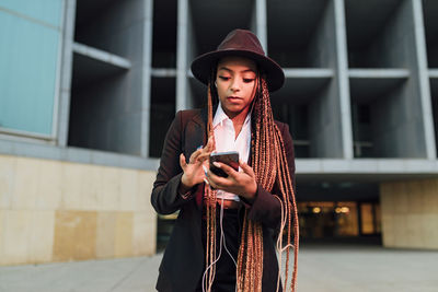 Businesswoman with smart phone listening music through in-ear headphones outside office building