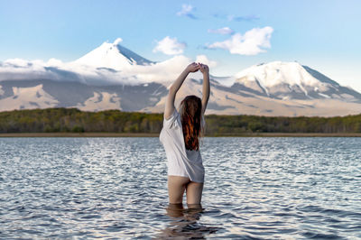 Rear view of woman with arms raised standing in lake against snowcapped mountains