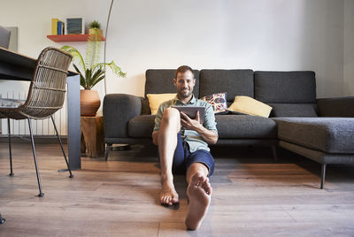 Smiling man sitting on floor with digital tablet against sofa at home