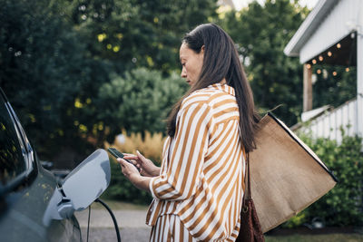 Focused woman carrying bags and using smart phone while charging car in back yard