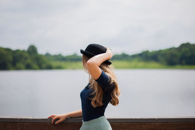 Woman wearing bowler hat standing by railing against lake