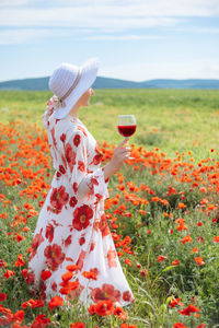 Rear view of a woman in summer dress with a glass of red wine among the field with blooming poppies