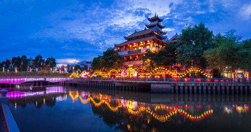 Illuminated building by lake against sky at dusk