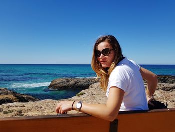 Portrait of woman wearing sunglasses sitting on bench at beach against clear sky