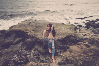 Woman listening music through headphones while sitting on rock at beach against sea
