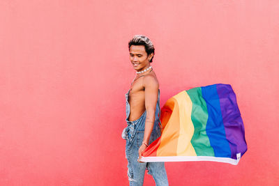 Side view of gay man holding rainbow flag against pink wall