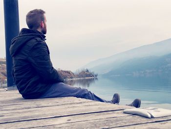 Side view of young man sitting on lake against sky