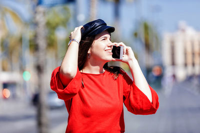 Smiling teenage girl wearing red top while using mobile phone in city during sunny day