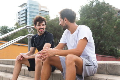 Low angle of cheerful hispanic sportsmen smiling and looking at each other while sitting on steps and speaking during break in fitness workout on street