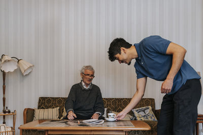 Smiling male care assistant serving tea to senior man sitting on sofa at home