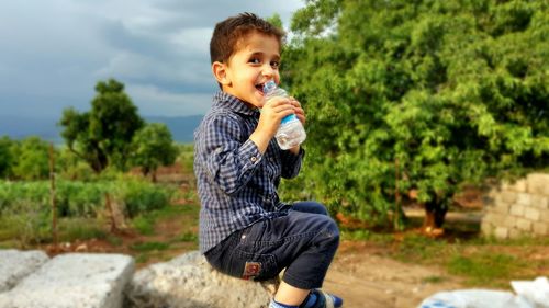 Portrait of smiling boy drinking water from bottle while sitting on rock