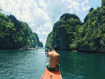 Rear view of shirtless man sitting in boat on sea against cloudy sky