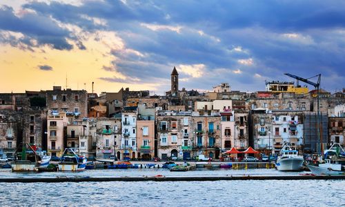 Cityscape by sea against cloudy sky during sunset