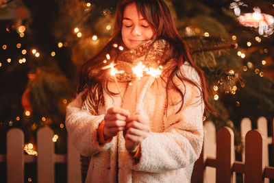 Happy teenage girl holding sparklers standing by christmas tree