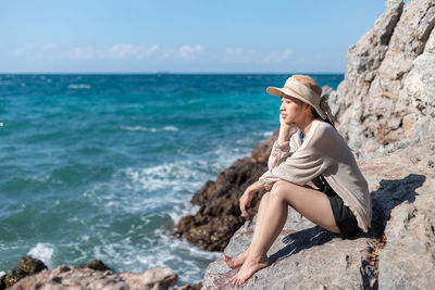 Full length of woman sitting on rock at beach against sky