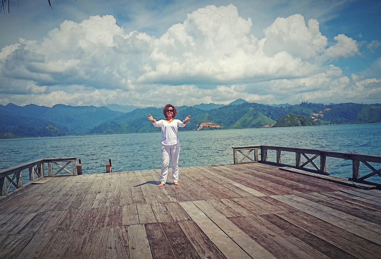 water, sky, full length, mountain, lifestyles, railing, leisure activity, cloud - sky, standing, casual clothing, rear view, tranquil scene, tranquility, mountain range, pier, sea, scenics, beauty in nature