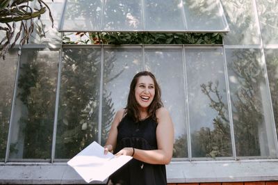 Young woman laughing while standing by greenhouse