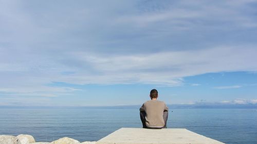 Rear view of man sitting on pier in sea against cloudy sky