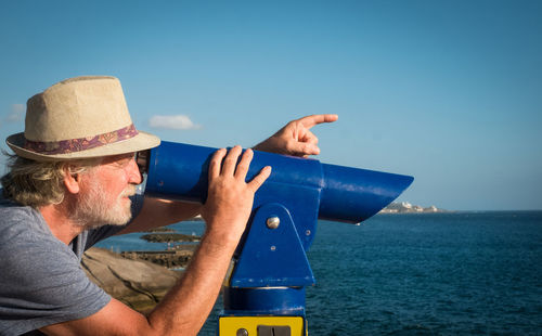 Man wearing hat looking through coin-operated telescope by sea against blue sky