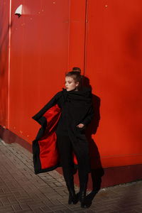 Fashionable teenage girl wearing black warm clothing standing against red wall during sunny day