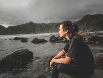 Side view of young woman standing on rock by sea against sky