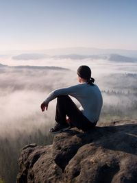 Man sit on the peak of rock and watching into colorful mist and fog in forest valley.