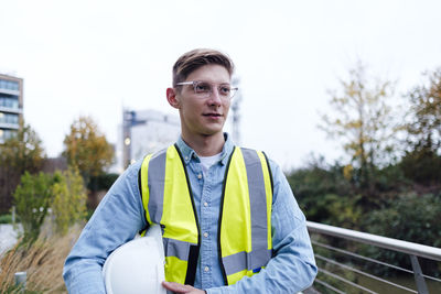 Contemplative engineer wearing eyeglasses holding hardhat under arm