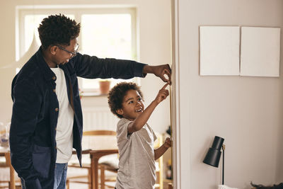 Happy father and son checking height while standing near wall at home