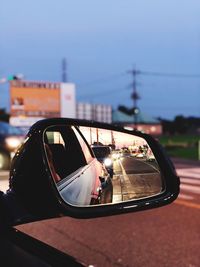 Reflection of cars in side-view mirror against sky at dusk