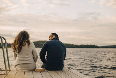 Rear view of couple talking while sitting on jetty by lake against sky
