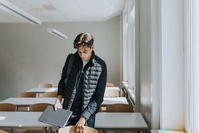 Teenage boy holding laptop while standing in classroom