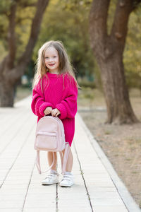 Funny cute kid girl 5-6 year old wear pink knit sweater holding backpack standing on road in park