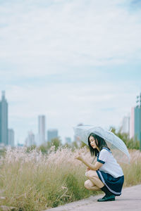 Side view of young woman with umbrella crouching by plants on field