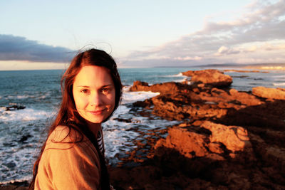 Portrait of smiling young woman sitting at beach against sky during sunset