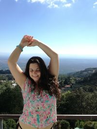 Smiling woman with arms raised standing against sky