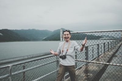 Full length of smiling young woman standing on railing against sky