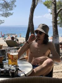 Fron view of a young man sitting at beach drinking beer against sky