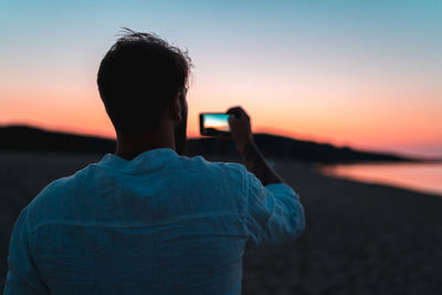 Rear view of man photographing sea against sky during sunset